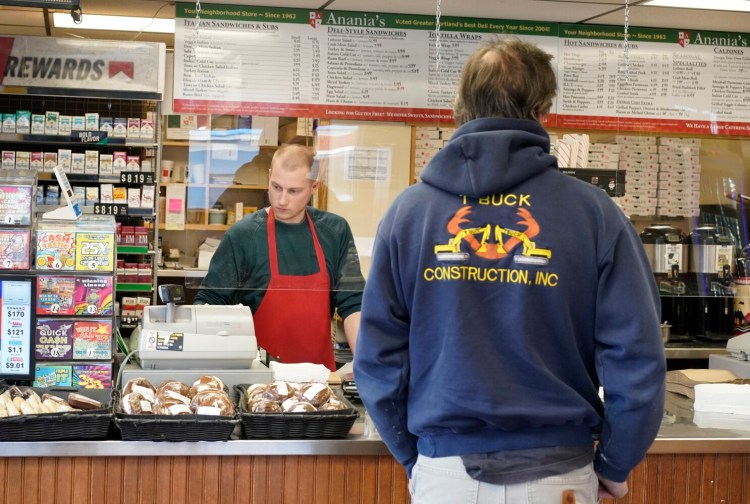 Behind a Plexiglas sheet hanging from the ceiling, Pete Hill helps a customer at Anania's in Portland on Wednesday. The store added the Plexiglas sheets at their registers to shield cashiers from customers.