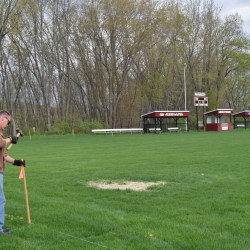 In the foreground, a man in a brown sweatshirt and blue jeans pounds a mallet into a wooden pole. He stands on a grassy field with a patch covered in tan hay. In the background, there is an electronic scoreboard and two shelters that have "University of Maine Farmington" logos on them.