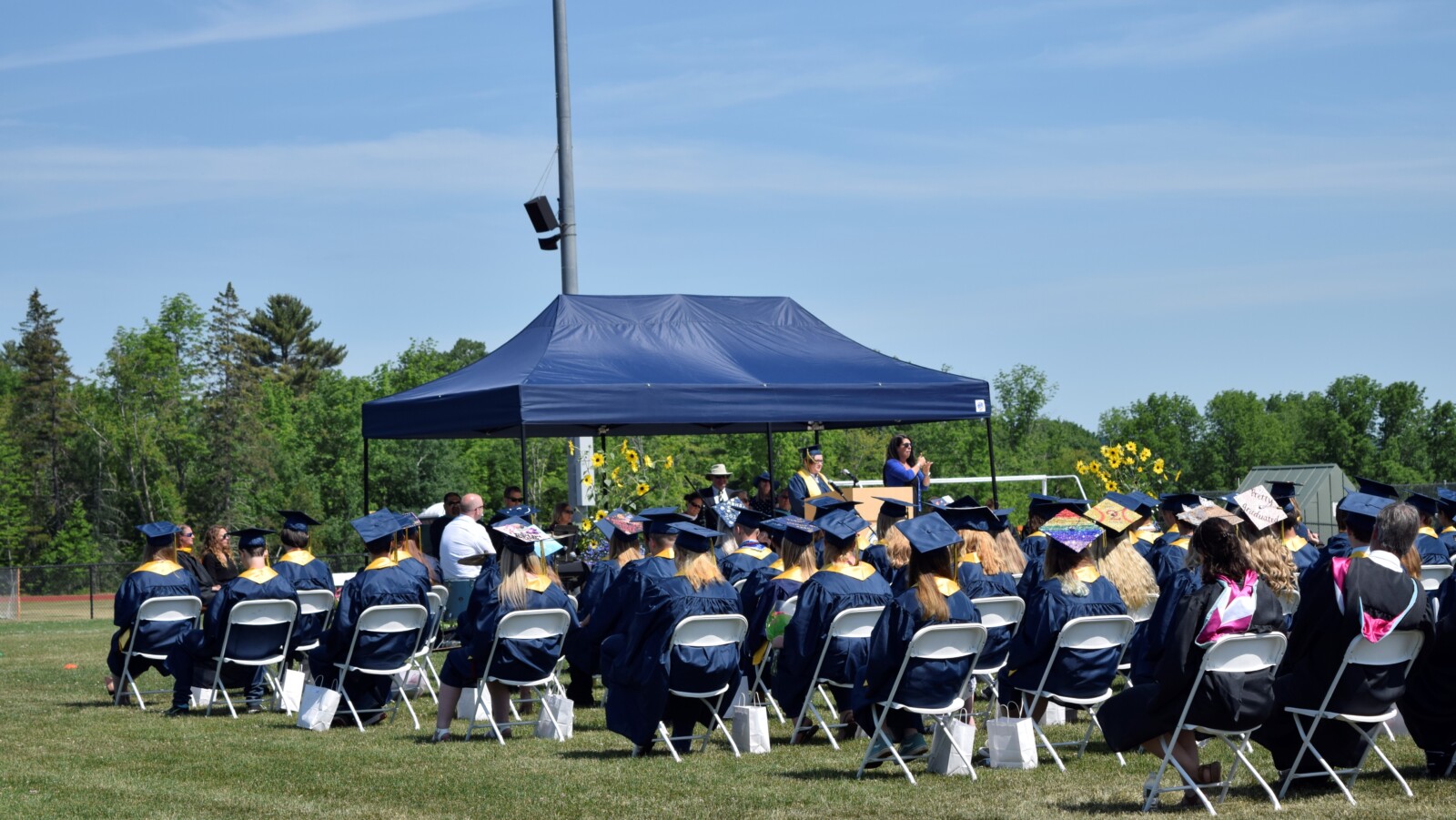 In the foreground, high school graduates sit in blue robes and caps. Some of the caps are decorated with phrases, LGBTQ rainbow-flag colors, and personal pictures. In the background, a student in a robe and cap stands on stage speaking into a microphone. Next to the student is an ASL interpreter.